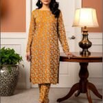 Model standing by a table lamp wearing the mustard orange variant of the ivy bunch 2 piece premium summer lawn suit with cream foliage prints.